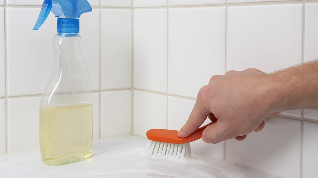 Nettoyage des joints de salle de bain : main brossant un joint de carrelage avec mousse, spray de produit à côté, carrelage blanc, scène réaliste et lumineuse.
